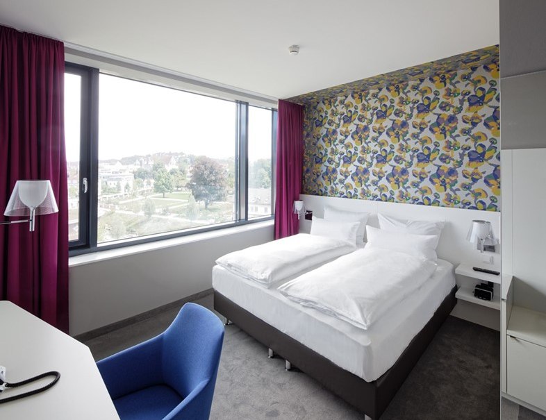 Modernes Hotelzimmer mit Doppelbett, bunter Tapete, großem Fenster und lila Vorhängen. Ein blauer Stuhl steht am Schreibtisch., © Hotel am Remspark Modernes Hotelzimmer mit Doppelbett, bunter Tapete, großem Fenster und lila Vorhängen. Ein blauer Stuhl steht am Schreibtisch., © Hotel am Remspark