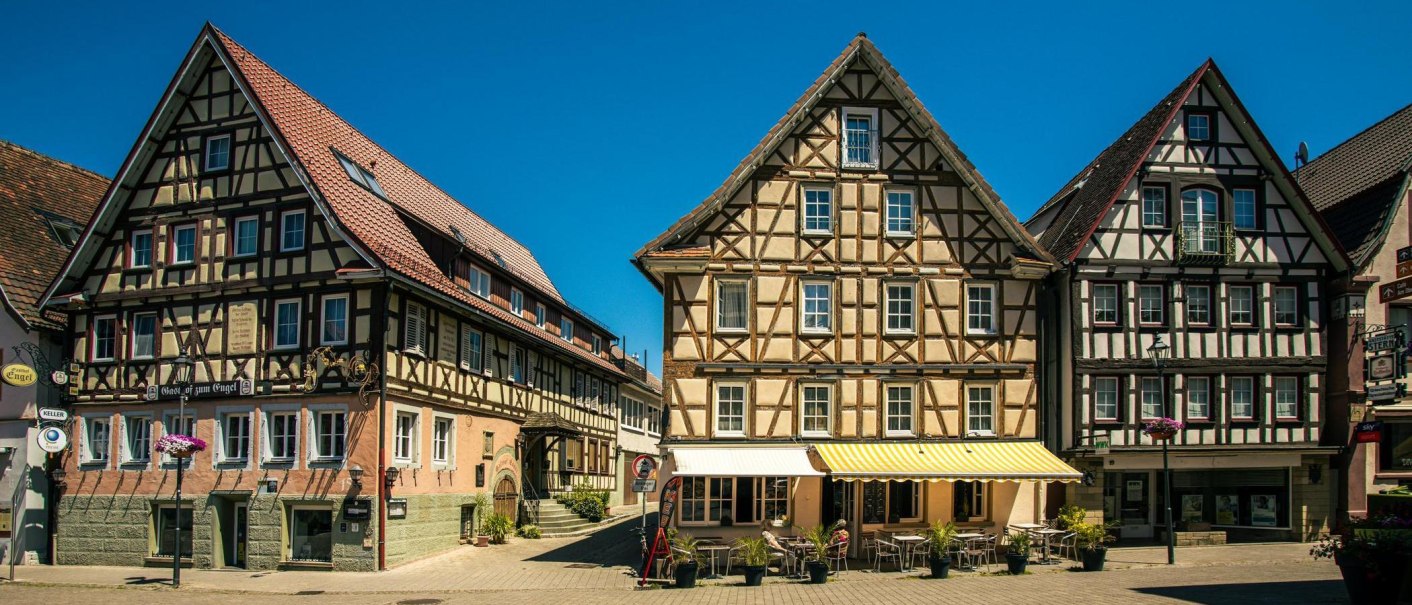 Fachwerkhäuser am Marktplatz von Murrhardt unter klarem, blauem Himmel. Vor den Gebäuden stehen Tische und Stühle eines Cafés., © SMG, Sarah Schmid Fachwerkhäuser am Marktplatz von Murrhardt unter klarem, blauem Himmel. Vor den Gebäuden stehen Tische und Stühle eines Cafés., © SMG, Sarah Schmid