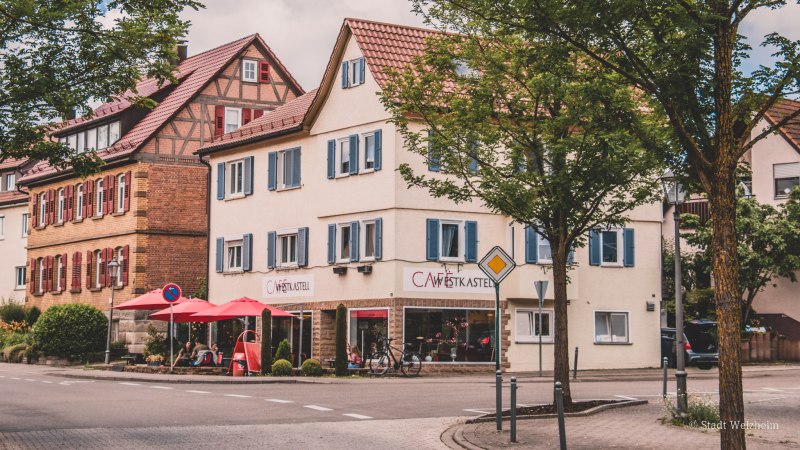 Ein Fachwerkhaus und ein Caf&eacute; mit roten Sonnenschirmen an einer Stra&szlig;enecke in einer deutschen Stadt, umgeben von B&auml;umen., &copy; Stadt Welzheim