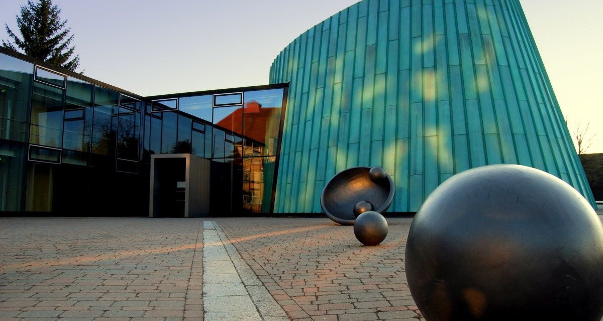 Moderne Architektur der Musikschule Fellbach mit Glasfassade und grüner Metallverkleidung. Im Vordergrund sind schwarze Kugelskulpturen zu sehen., © Simone Mathias Moderne Architektur der Musikschule Fellbach mit Glasfassade und grüner Metallverkleidung. Im Vordergrund sind schwarze Kugelskulpturen zu sehen., © Simone Mathias