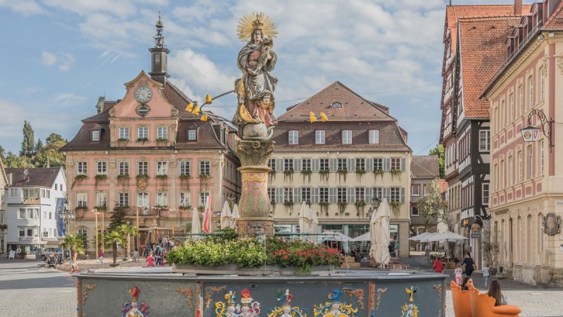 Ein belebter Marktplatz mit einem verzierten Brunnen und einem historischen Rathaus im Hintergrund. Menschen sitzen auf orangen St&uuml;hlen.