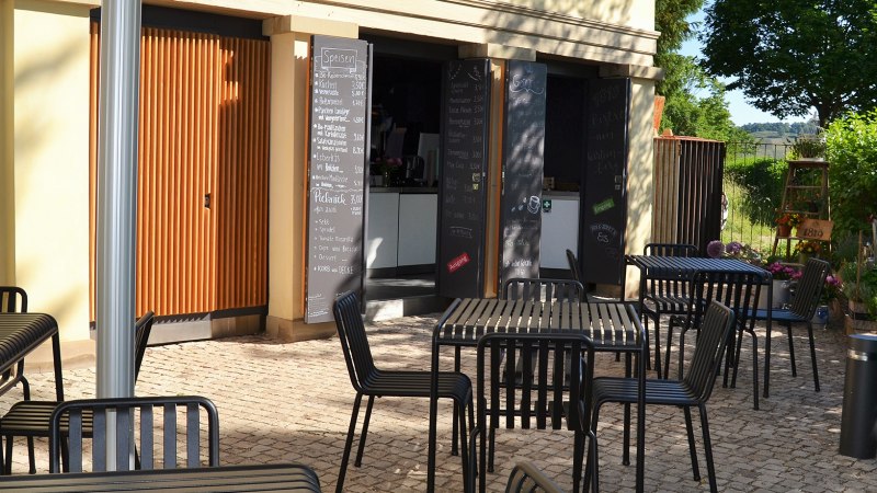 Außenbereich des 1819 Bistros am Wirtemberg mit schwarzen Tischen und Stühlen auf gepflastertem Boden, umgeben von grüner Vegetation., © 1819 Bistro am Wirtemberg