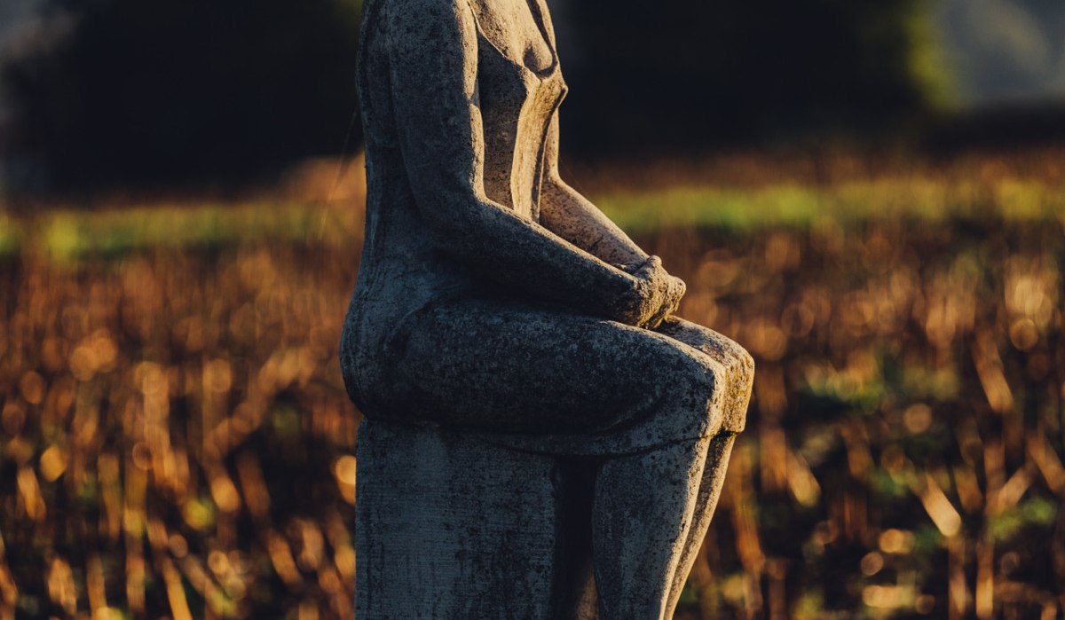 Steinskulptur einer sitzenden Frau auf einem Feld, beleuchtet von warmem Sonnenlicht. Im Hintergrund sind Bäume und ein Hügel zu sehen., © Landratsamt Ostalbkreis
