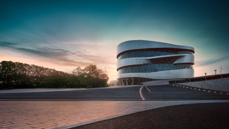 Das Mercedes-Benz Museum in Stuttgart bei Sonnenuntergang. Moderne Architektur mit geschwungenen Linien, umgeben von Bäumen und beleuchteten Stufen., © Mercedes-Benz Heritage GmbH Das Mercedes-Benz Museum in Stuttgart bei Sonnenuntergang. Moderne Architektur mit geschwungenen Linien, umgeben von Bäumen und beleuchteten Stufen., © Mercedes-Benz Heritage GmbH