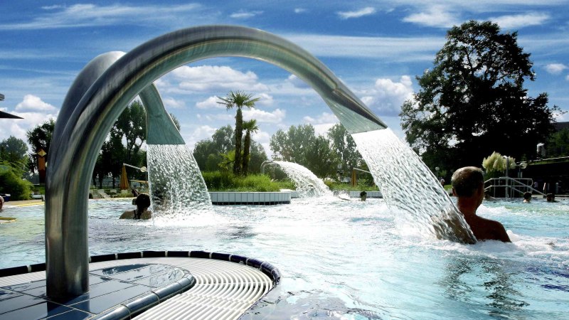 Ein Außenpool mit Wasserfontänen und Palmen, umgeben von Bäumen und einem Gebäude. Menschen genießen das Wasser unter blauem Himmel., © Stuttgarter Bäder