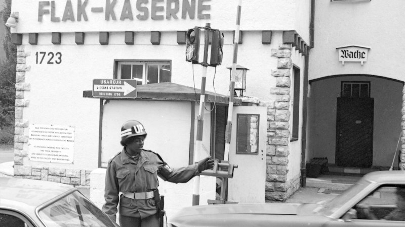 Ein Soldat in Uniform dirigiert den Verkehr vor der Flak-Kaserne. Ein Schild weist auf die USAREUR License Station hin. Autos fahren vorbei., © Ludwigsburg Museum Ein Soldat in Uniform dirigiert den Verkehr vor der Flak-Kaserne. Ein Schild weist auf die USAREUR License Station hin. Autos fahren vorbei., © Ludwigsburg Museum