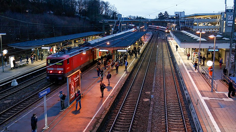 Bahnhof Backnang bei Dämmerung. Ein roter Zug steht am Gleis, Menschen warten auf den Bahnsteigen. Beleuchtung und Schienen sind sichtbar., © Alex Becher
