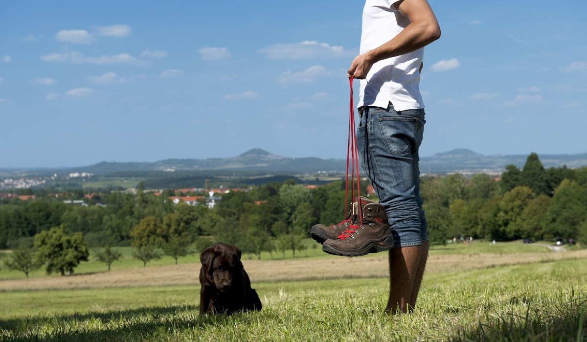 Person in Jeans und weißem T-Shirt hält Wanderschuhe. Ein Hund sitzt im Gras. Im Hintergrund sind grüne Felder und Hügel zu sehen., © Landkreis Göppingen Person in Jeans und weißem T-Shirt hält Wanderschuhe. Ein Hund sitzt im Gras. Im Hintergrund sind grüne Felder und Hügel zu sehen., © Landkreis Göppingen