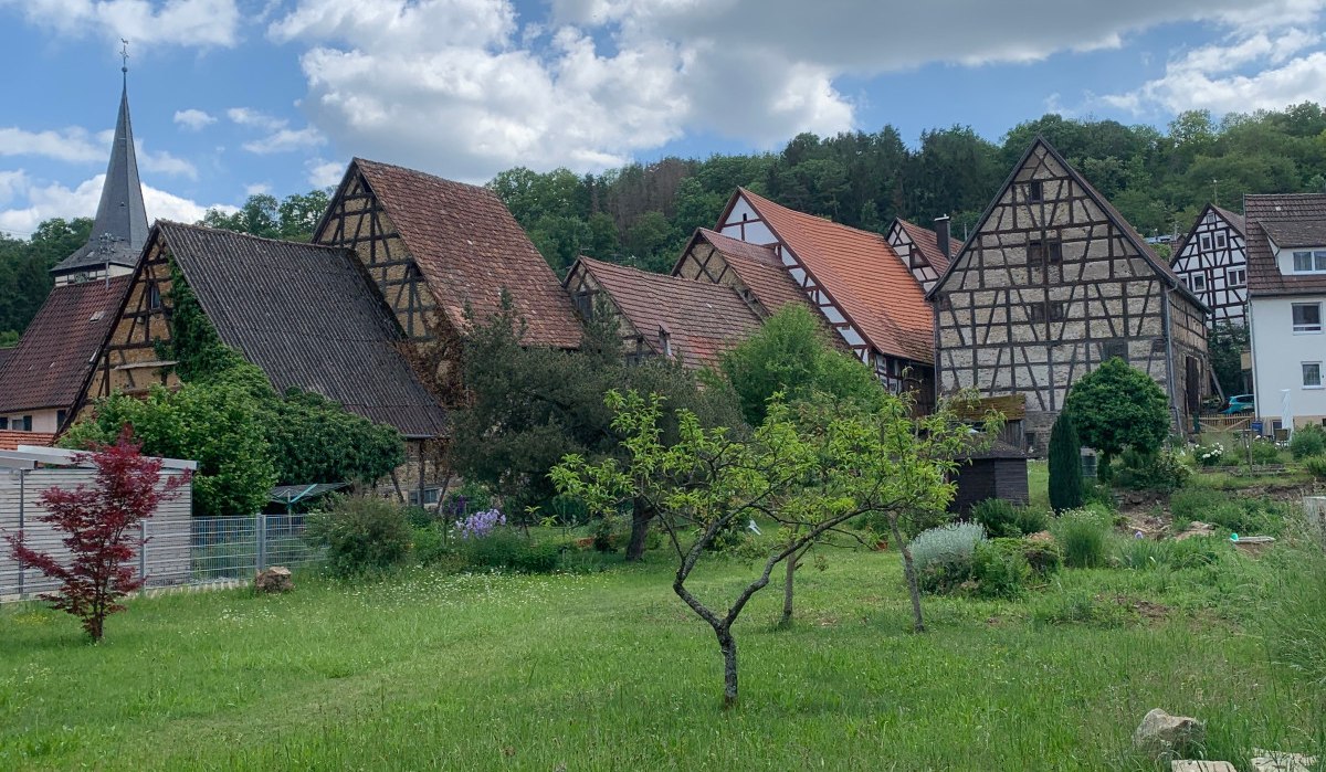 Fachwerkhäuser mit roten Dächern und ein Kirchturm in einem Dorf, umgeben von grüner Landschaft und Bäumen., © Land der 1000 Hügel - Kraichgau-Stromberg Fachwerkhäuser mit roten Dächern und ein Kirchturm in einem Dorf, umgeben von grüner Landschaft und Bäumen., © Land der 1000 Hügel - Kraichgau-Stromberg