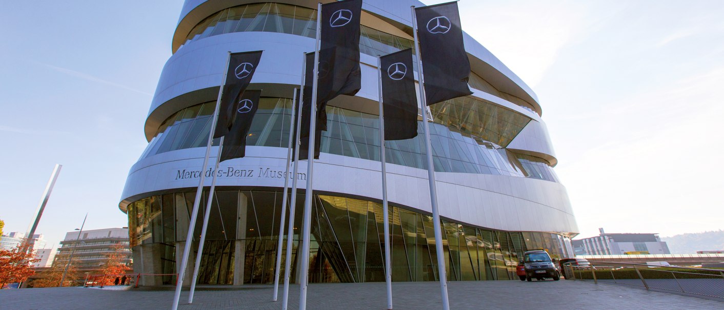 Das Mercedes-Benz Museum mit seiner modernen Architektur und mehreren Fahnen im Vordergrund. Der Himmel ist klar und blau., © SMG, Achim Mende