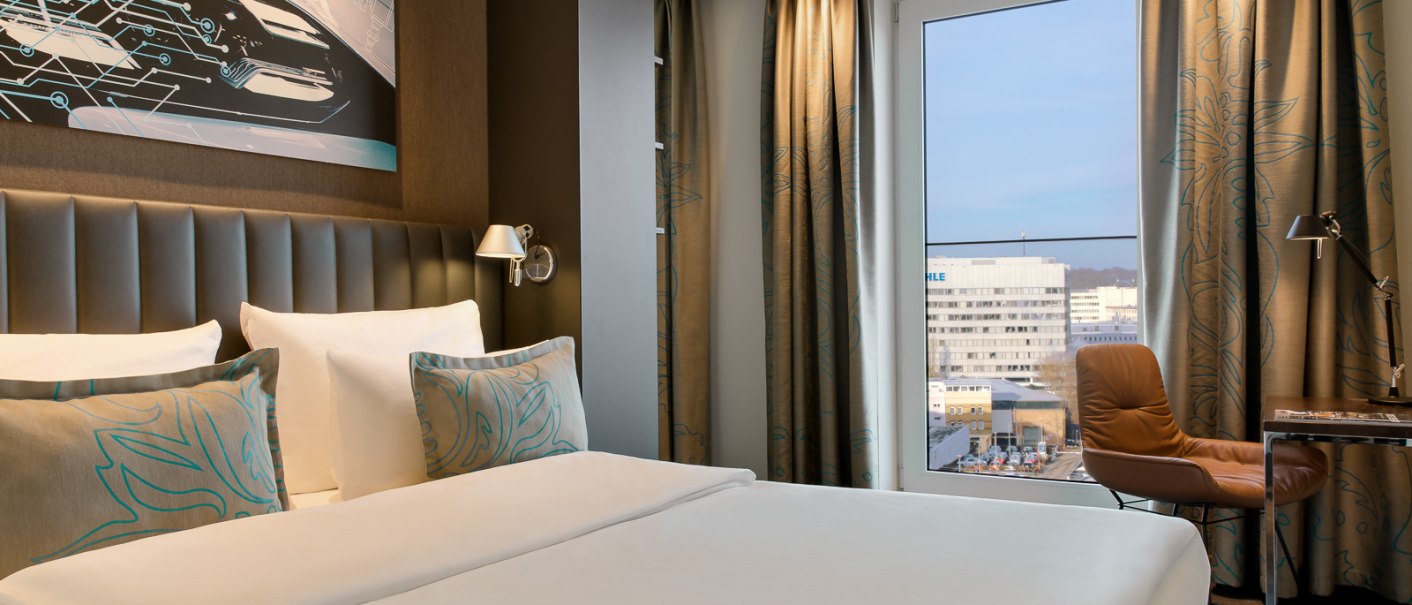 Modernes Hotelzimmer mit großem Bett, Kunstwerk an der Wand, Fenster mit Stadtblick, braunem Sessel und Schreibtisch., © TOMAS