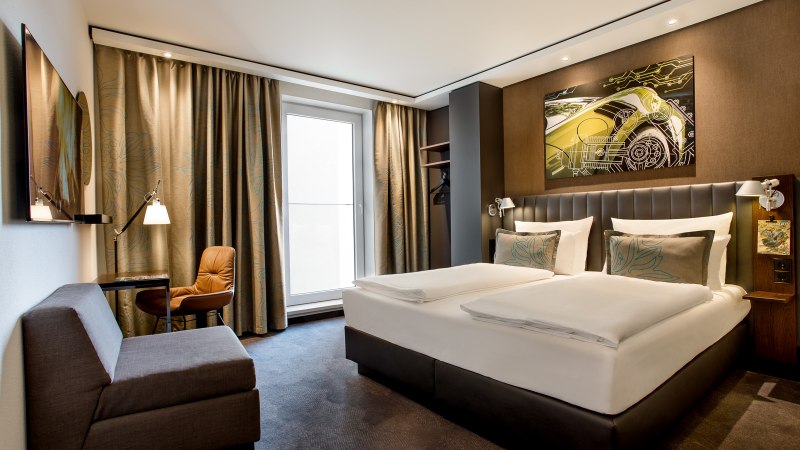 Modernes Hotelzimmer mit Doppelbett, Sofa, Schreibtisch und großem Fenster. An der Wand hängt ein Kunstwerk mit einem Auto-Motiv., © TOMAS Modernes Hotelzimmer mit Doppelbett, Sofa, Schreibtisch und großem Fenster. An der Wand hängt ein Kunstwerk mit einem Auto-Motiv., © TOMAS