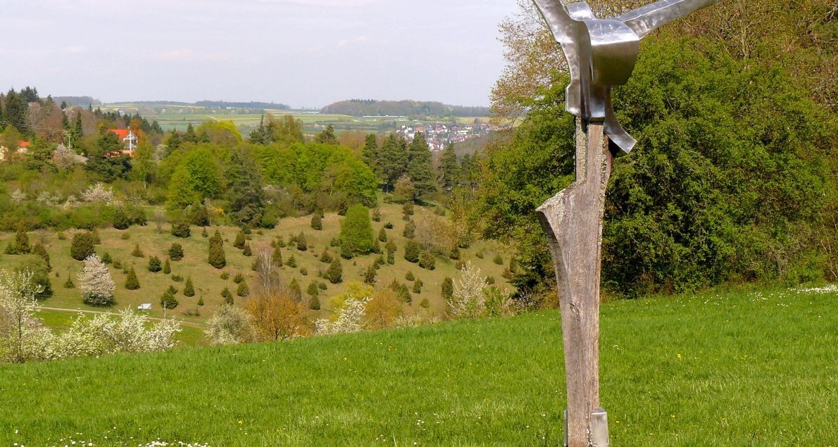 Eine moderne Skulptur steht auf einer grünen Wiese mit Blick auf eine hügelige Landschaft und blühende Bäume., © Natur.Nah. Schönbuch & Heckengäu Eine moderne Skulptur steht auf einer grünen Wiese mit Blick auf eine hügelige Landschaft und blühende Bäume., © Natur.Nah. Schönbuch & Heckengäu