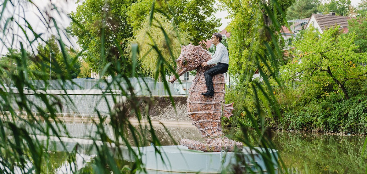 Ein Mann reitet auf einer großen Seepferdchen-Skulptur im Wasser, umgeben von grünen Bäumen und Pflanzen., © Unbekannt