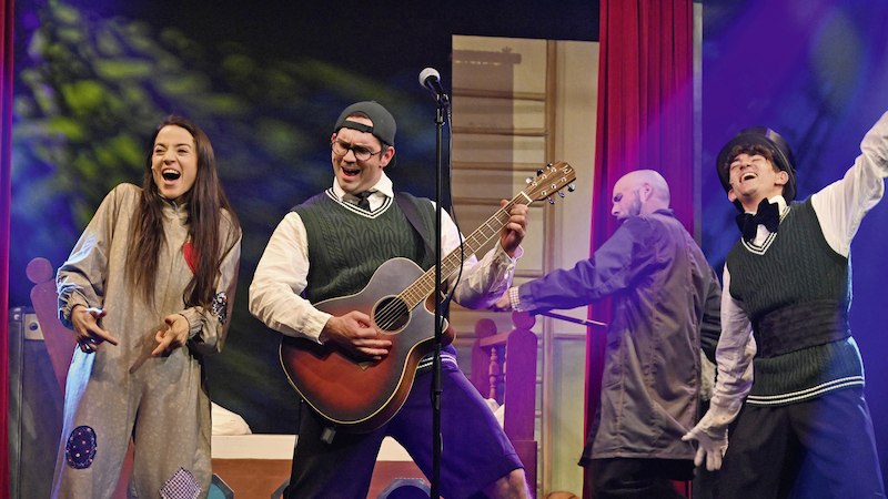 Vier Darsteller auf einer Bühne, einer spielt Gitarre, die anderen tanzen und lachen. Bunte Lichter und Requisiten im Hintergrund., © Stadtverwaltung Schorndorf