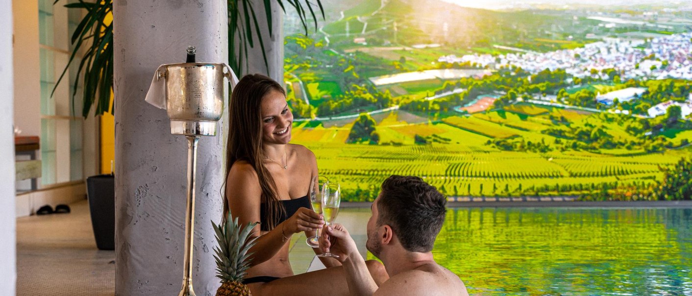 Ein Paar sitzt am Pool, trinkt Champagner und lächelt sich an. Im Hintergrund ist ein großes Landschaftsbild zu sehen., © Ringhotel Adler Asperg