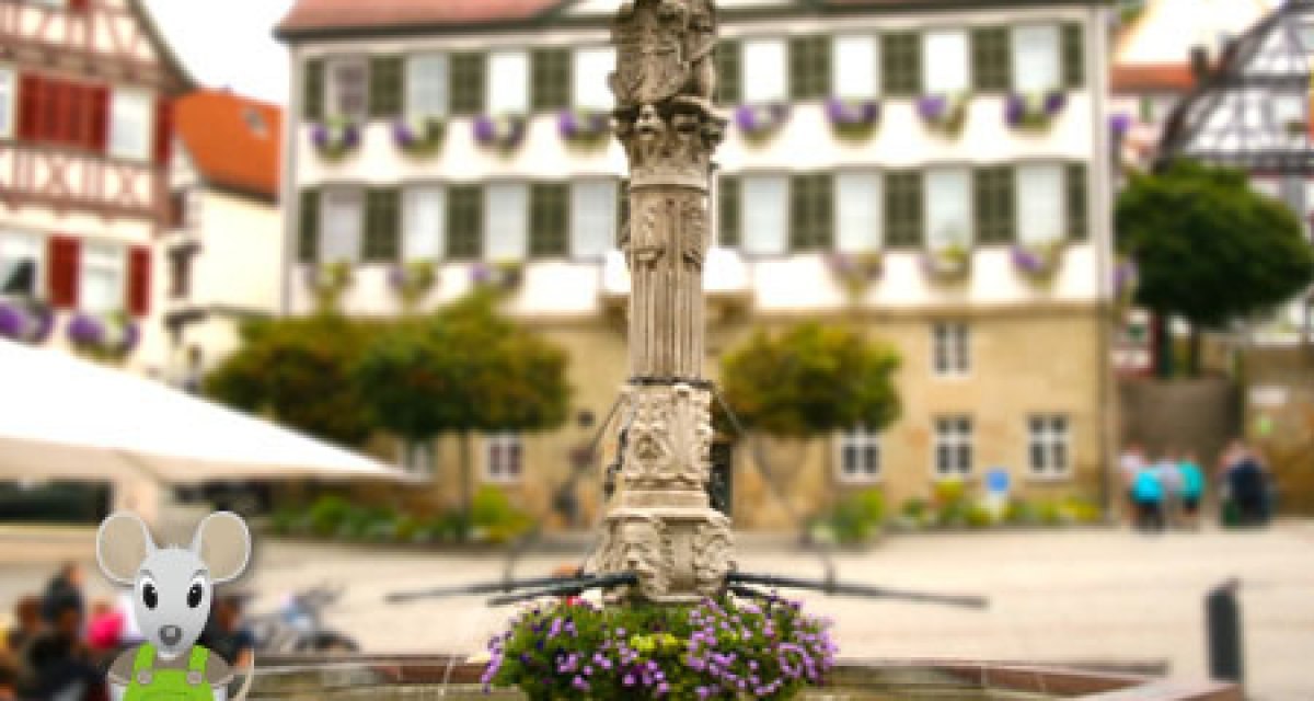 Ein verzierter Marktbrunnen steht im Zentrum eines Platzes, umgeben von Fachwerkhäusern. Eine Zeichentrickmaus ist im Vordergrund zu sehen. Ein verzierter Marktbrunnen steht im Zentrum eines Platzes, umgeben von Fachwerkhäusern. Eine Zeichentrickmaus ist im Vordergrund zu sehen.