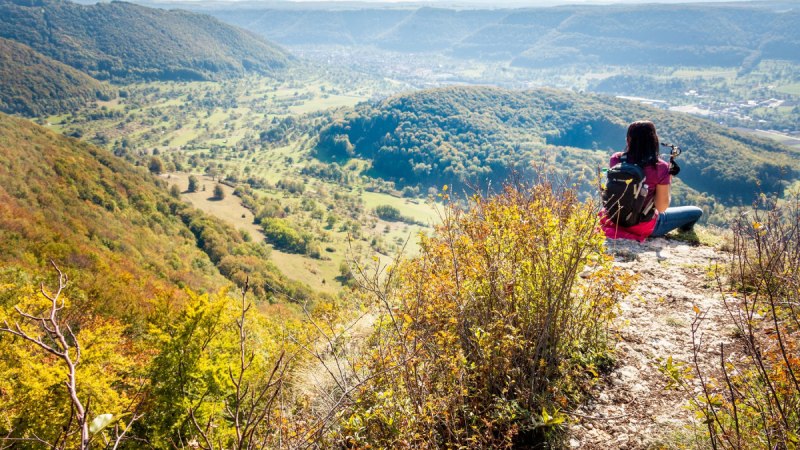 Eine Person sitzt auf einem Hügel und blickt ins Lenninger Tal. Die Landschaft ist von bewaldeten Hügeln und weiten Wiesen geprägt., © hochgehberge