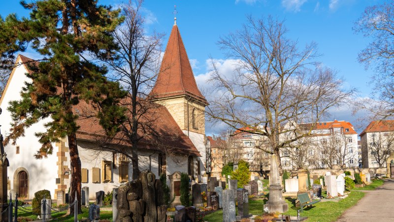Der Uff-Kirchhof mit Kirche, Grabsteinen und B&auml;umen unter blauem Himmel; im Hintergrund stehen Wohnh&auml;user., &copy; Peter Jagusch/Landeshauptstadt Stuttgart