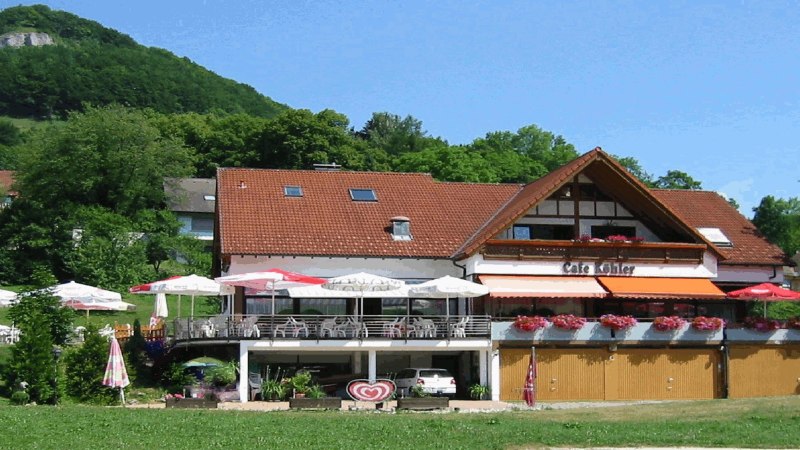 Café Köhler mit Terrasse und Sonnenschirmen, umgeben von grüner Landschaft und einem bewaldeten Hügel im Hintergrund., © Café Köhler Café Köhler mit Terrasse und Sonnenschirmen, umgeben von grüner Landschaft und einem bewaldeten Hügel im Hintergrund., © Café Köhler