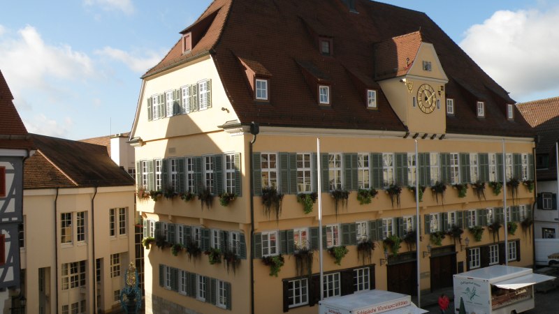 Das historische Rathaus von Nürtingen mit gelber Fassade und grünen Fensterläden. Vor dem Gebäude sind Verkaufsstände und Fahrräder zu sehen., © Schreitmüller Das historische Rathaus von Nürtingen mit gelber Fassade und grünen Fensterläden. Vor dem Gebäude sind Verkaufsstände und Fahrräder zu sehen., © Schreitmüller