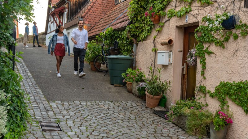 Ein Paar spaziert durch eine malerische Altstadtstraße in Besigheim. Fachwerkhäuser und Pflanzen schmücken den Weg. Weitere Personen im Hintergrund., © SMG, Martina Denker Ein Paar spaziert durch eine malerische Altstadtstraße in Besigheim. Fachwerkhäuser und Pflanzen schmücken den Weg. Weitere Personen im Hintergrund., © SMG, Martina Denker