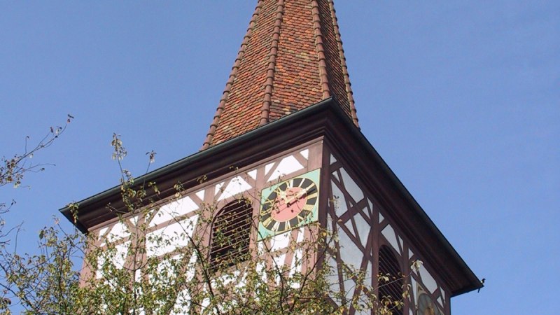 Ein Kirchturm mit Fachwerk und Uhr ragt über Bäume in den blauen Himmel. Der Turm hat ein spitzes Dach mit Wetterhahn., © Natur.Nah. Schönbuch & Heckengäu