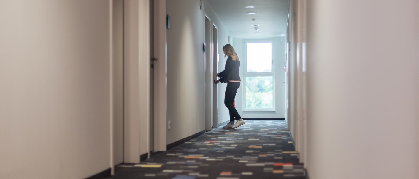 Eine Person steht in einem langen Flur und öffnet eine Tür. Der Flur ist hell und hat einen bunten Teppichboden., © the niu Form - Novum Hospitality