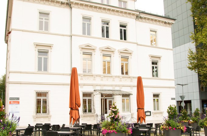 Historisches Gebäude mit weißer Fassade, davor ein Café mit Tischen und Pflanzen. Die Architektur erinnert an die frühere Weberei Gutmann., © Stadt Göppingen Historisches Gebäude mit weißer Fassade, davor ein Café mit Tischen und Pflanzen. Die Architektur erinnert an die frühere Weberei Gutmann., © Stadt Göppingen