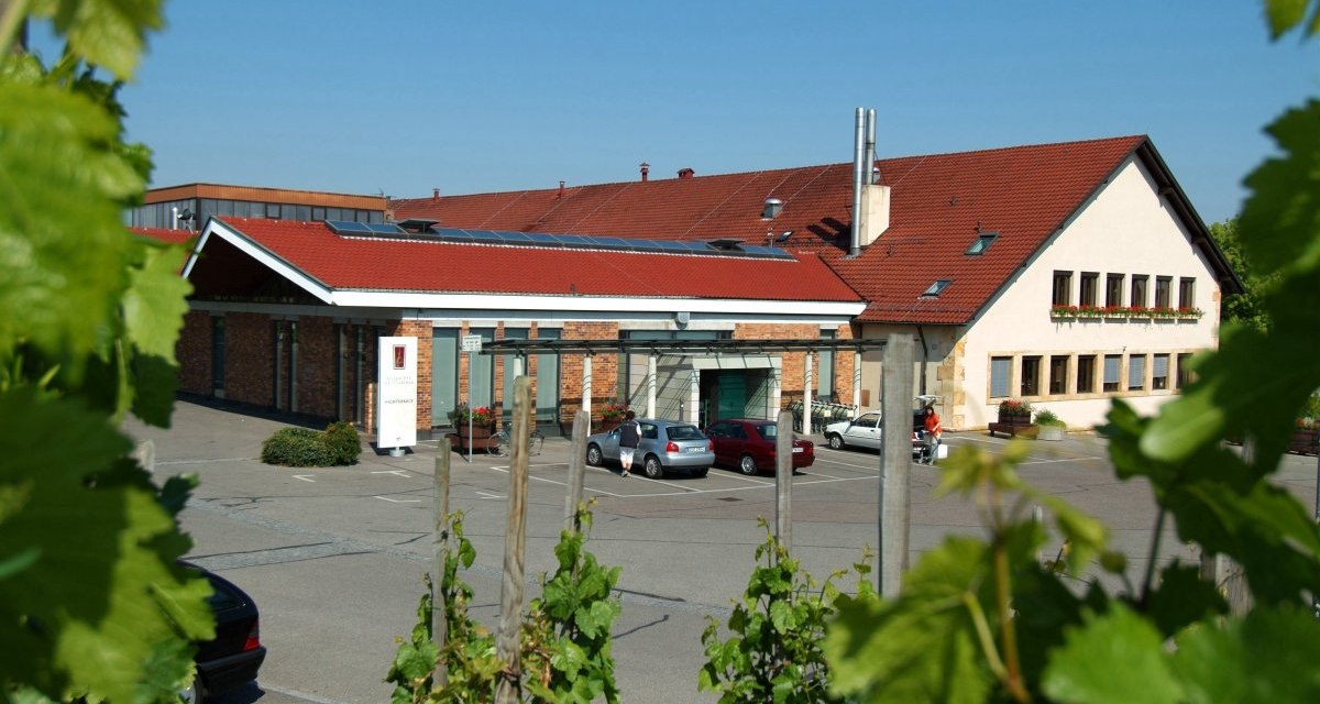 Gebäude der Fellbacher Weingärtner mit rotem Dach, umgeben von Weinreben. Autos parken auf dem Vorplatz. Klare, sonnige Wetterbedingungen., © Remstal Tourismus e.V. Gebäude der Fellbacher Weingärtner mit rotem Dach, umgeben von Weinreben. Autos parken auf dem Vorplatz. Klare, sonnige Wetterbedingungen., © Remstal Tourismus e.V.