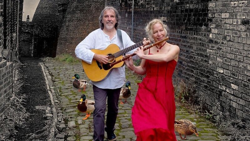 Zwei Musiker, eine Frau in rotem Kleid mit Flöte und ein Mann mit Gitarre, spazieren auf einem Kopfsteinpflasterweg. Enten und alte Schornsteine im Hintergrund., © Mauerwerk GmbH