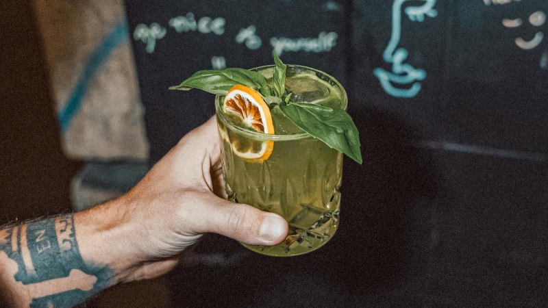 Eine tätowierte Hand hält ein grünes Getränk mit Basilikum und Orangenscheibe. Im Hintergrund ist eine Tafel mit Zeichnungen und Schrift zu sehen., © Bruno, Stuttgart Eine tätowierte Hand hält ein grünes Getränk mit Basilikum und Orangenscheibe. Im Hintergrund ist eine Tafel mit Zeichnungen und Schrift zu sehen., © Bruno, Stuttgart