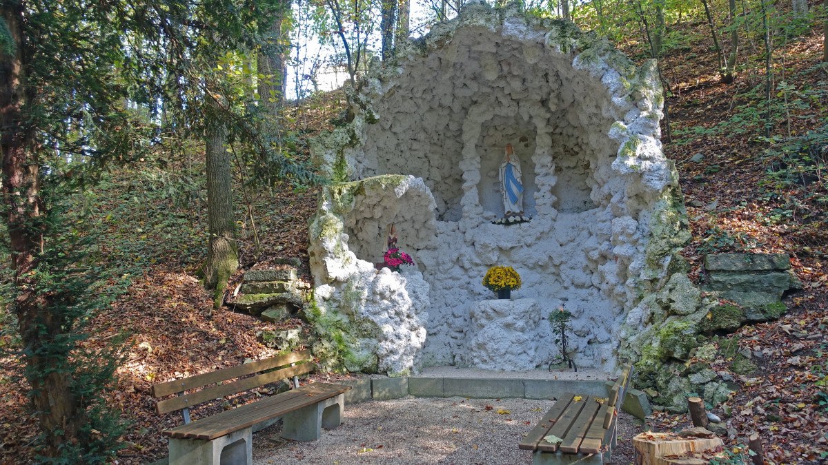 Die Lourdesgrotte in Böbingen zeigt eine Marienstatue in einer steinernen Nische, umgeben von Wald. Vor der Grotte stehen Holzbänke., © Foto: Cornelia Steinbach