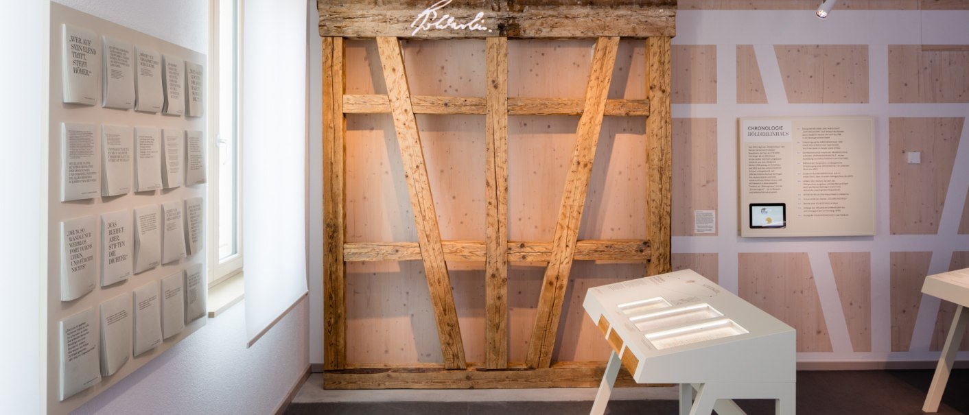 Ausstellungsraum mit Holzwand, Texttafeln und interaktiven Stationen. Ein Fenster links, beleuchtete Tafeln und ein Bildschirm rechts., © dieargelola, Stuttgart