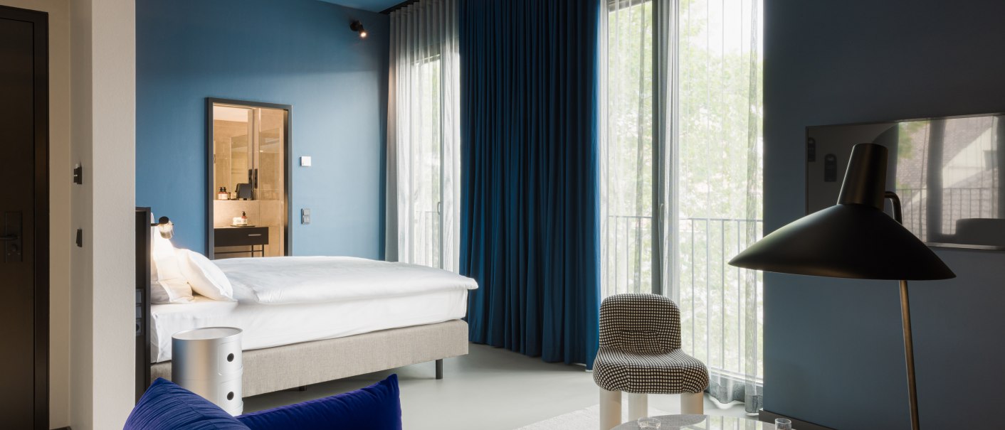 Modernes Hotelzimmer mit blauem Sofa, Bett, Tisch und Lampe. Große Fenster mit blauen Vorhängen und hellem Teppich., © EmiLu Hotel GmbH Modernes Hotelzimmer mit blauem Sofa, Bett, Tisch und Lampe. Große Fenster mit blauen Vorhängen und hellem Teppich., © EmiLu Hotel GmbH