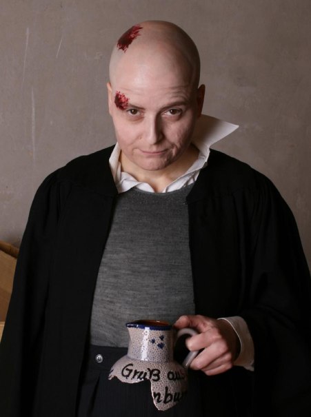 Eine Person mit Glatze und Kunstblut auf der Stirn h&auml;lt einen Krug mit der Aufschrift 'Gru&szlig; aus...'. Sie tr&auml;gt einen schwarzen Umhang und ein graues Oberteil., &copy; Foto_(c)_Dirk Holtkamp Endemann