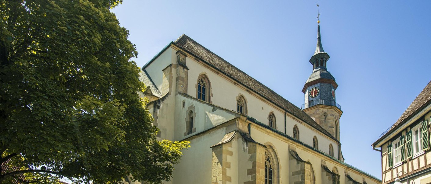 Historische Stadtkirche mit Turm und Uhr, umgeben von grünen Bäumen und Fachwerkhäusern unter blauem Himmel., © Stuttgart-Marketing GmbH, Sarah Schmid Historische Stadtkirche mit Turm und Uhr, umgeben von grünen Bäumen und Fachwerkhäusern unter blauem Himmel., © Stuttgart-Marketing GmbH, Sarah Schmid