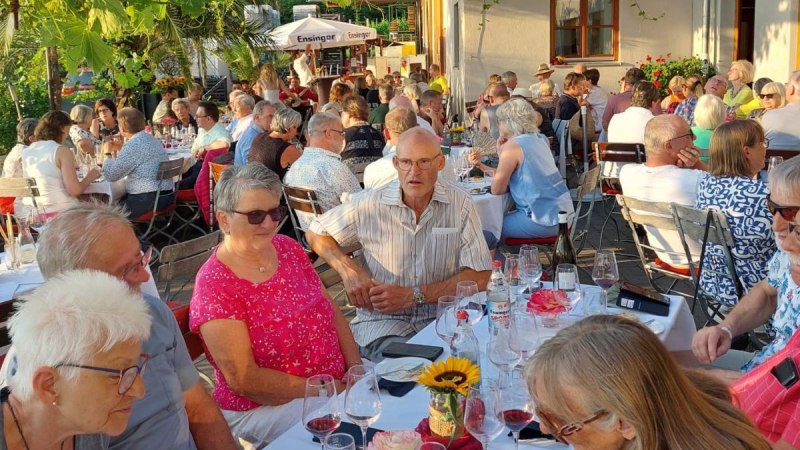 Geselliges Beisammensein im Freien unter Weinreben. Menschen sitzen an Tischen, genie&szlig;en Getr&auml;nke und unterhalten sich in entspannter Atmosph&auml;re., &copy; Weingut Bayer