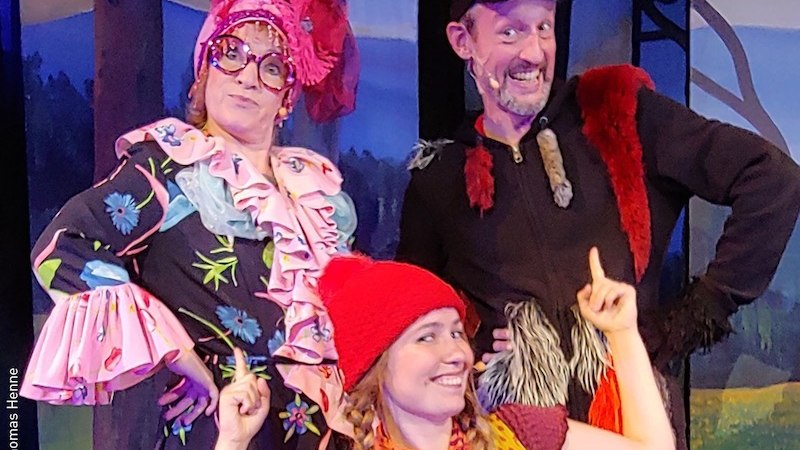 Drei Personen in bunten Kostümen auf einer Bühne. Eine Person trägt eine rote Mütze, die anderen fantasievolle Kleidung. Hintergrund zeigt eine Waldszene., © Stadt Kornwestheim Kulturmanagement