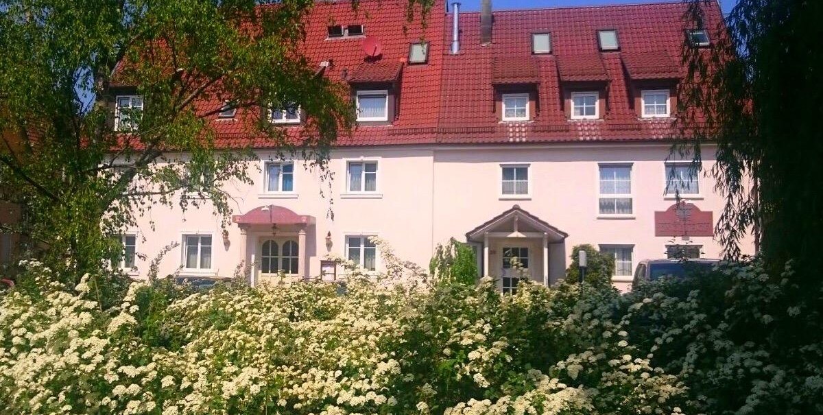 Ein Hotel mit rotem Dach und mehreren Fenstern, umgeben von blühenden Sträuchern und Bäumen, unter blauem Himmel., © Hotel Engelhof