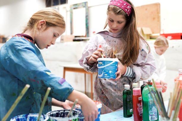 Zwei Kinder malen in einem Kunstworkshop. Sie tragen Malkittel und verwenden Pinsel und Farben. Im Hintergrund ist ein weiteres Kind zu sehen., © Kunstschule Labyrinth