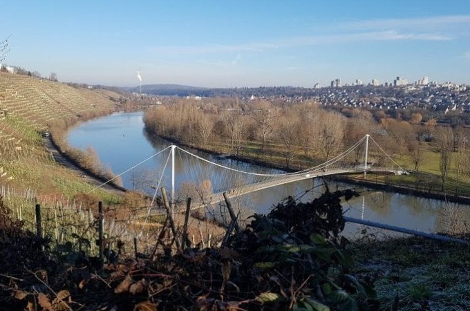 Hängebrücke über einen Fluss, umgeben von Weinbergen und kahlen Bäumen, mit einer Stadt im Hintergrund unter klarem Himmel., © Cool-Tours StattReisen Hängebrücke über einen Fluss, umgeben von Weinbergen und kahlen Bäumen, mit einer Stadt im Hintergrund unter klarem Himmel., © Cool-Tours StattReisen