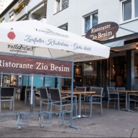 Außenbereich des Ristorante Zio Besim mit Tischen, Stühlen und einem großen Sonnenschirm. Das Restaurant hat eine moderne Glasfassade.