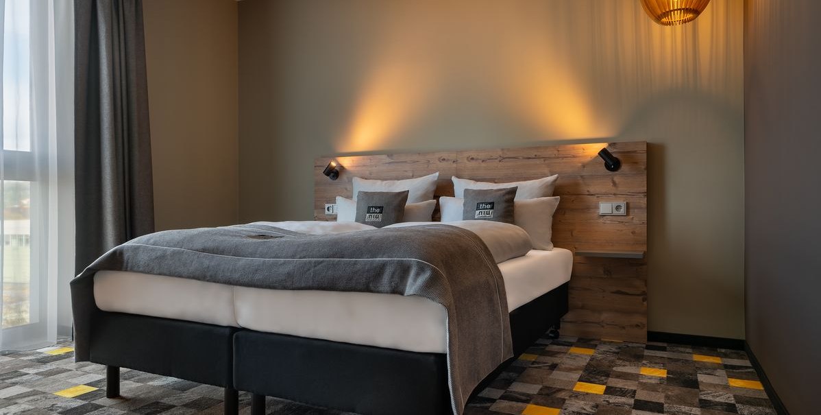Modernes Hotelzimmer mit Doppelbett, Holzkopfteil, grauer Bettwäsche und stilvoller Beleuchtung. Der Teppich hat ein geometrisches Muster., © Novum Hospitality Modernes Hotelzimmer mit Doppelbett, Holzkopfteil, grauer Bettwäsche und stilvoller Beleuchtung. Der Teppich hat ein geometrisches Muster., © Novum Hospitality