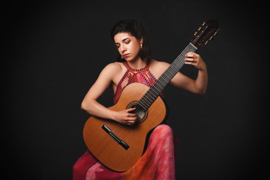 Eine Frau in einem bunten Kleid spielt Gitarre vor einem dunklen Hintergrund. Sie wirkt konzentriert und vertieft in die Musik.