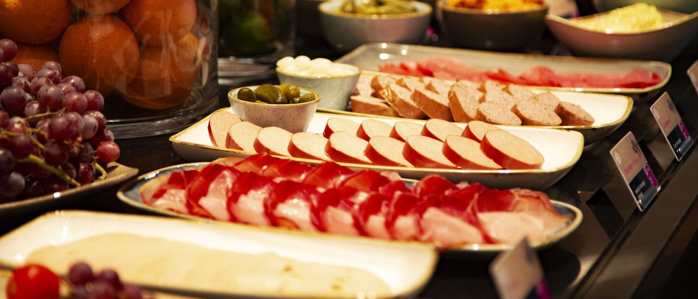 Ein Frühstücksbuffet mit verschiedenen Wurstsorten, Trauben, Orangen und eingelegten Gurken auf Tellern und Schalen arrangiert., © MASEVEN Stuttgart SI-Centrum Ein Frühstücksbuffet mit verschiedenen Wurstsorten, Trauben, Orangen und eingelegten Gurken auf Tellern und Schalen arrangiert., © MASEVEN Stuttgart SI-Centrum