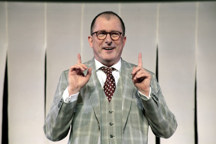 Ein Mann in einem karierten Anzug mit Krawatte gestikuliert und spricht auf einer Bühne., © Dietrich Dettmann