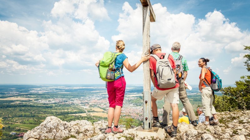 Wanderer stehen auf einem Berggipfel neben einem Gipfelkreuz und genießen die Aussicht auf die Landschaft unter einem blauen Himmel mit Wolken., © hochgehberge