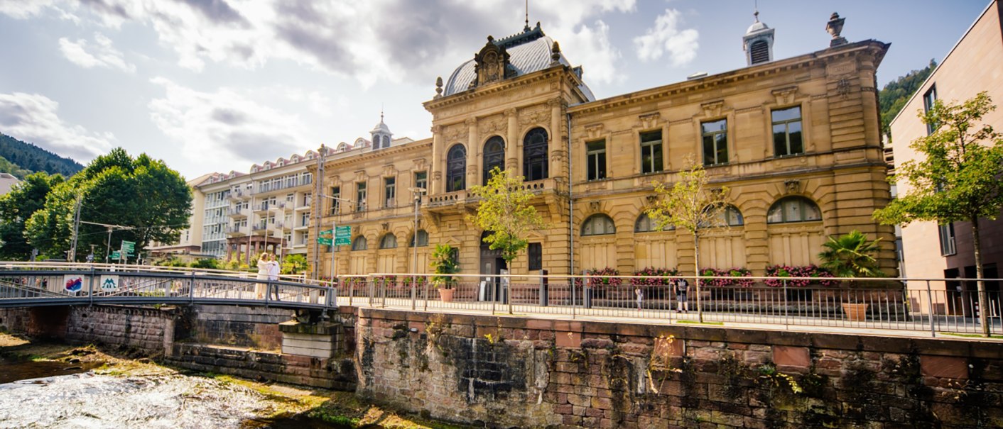 Das Forum König-Karls-Bad in Bad Wildbad, ein historisches Gebäude mit aufwendiger Fassade, steht neben einem Fluss und einer Brücke unter einem bewölkten Himmel., © Touristik Bad Wildbad GmbH