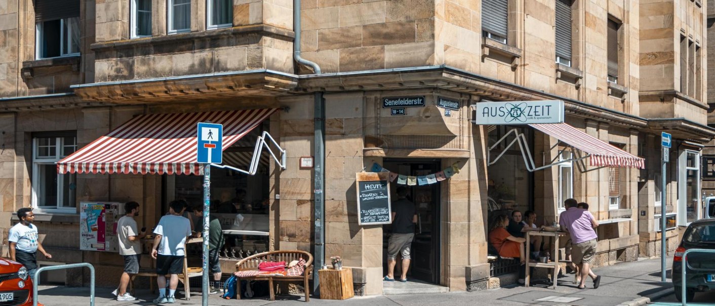 Cafébar Auszeit an einer Straßenecke mit roter Markise. Menschen sitzen draußen, Straßenschilder und Autos sind sichtbar., © SMG, Sarah Schmid Cafébar Auszeit an einer Straßenecke mit roter Markise. Menschen sitzen draußen, Straßenschilder und Autos sind sichtbar., © SMG, Sarah Schmid